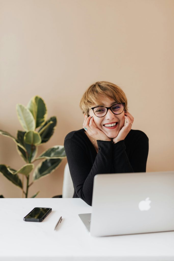 pexels-photo-4467687-4467687 Cheerful woman in eyeglasses and black sweater smiling while working at a desk with a laptop indoors.